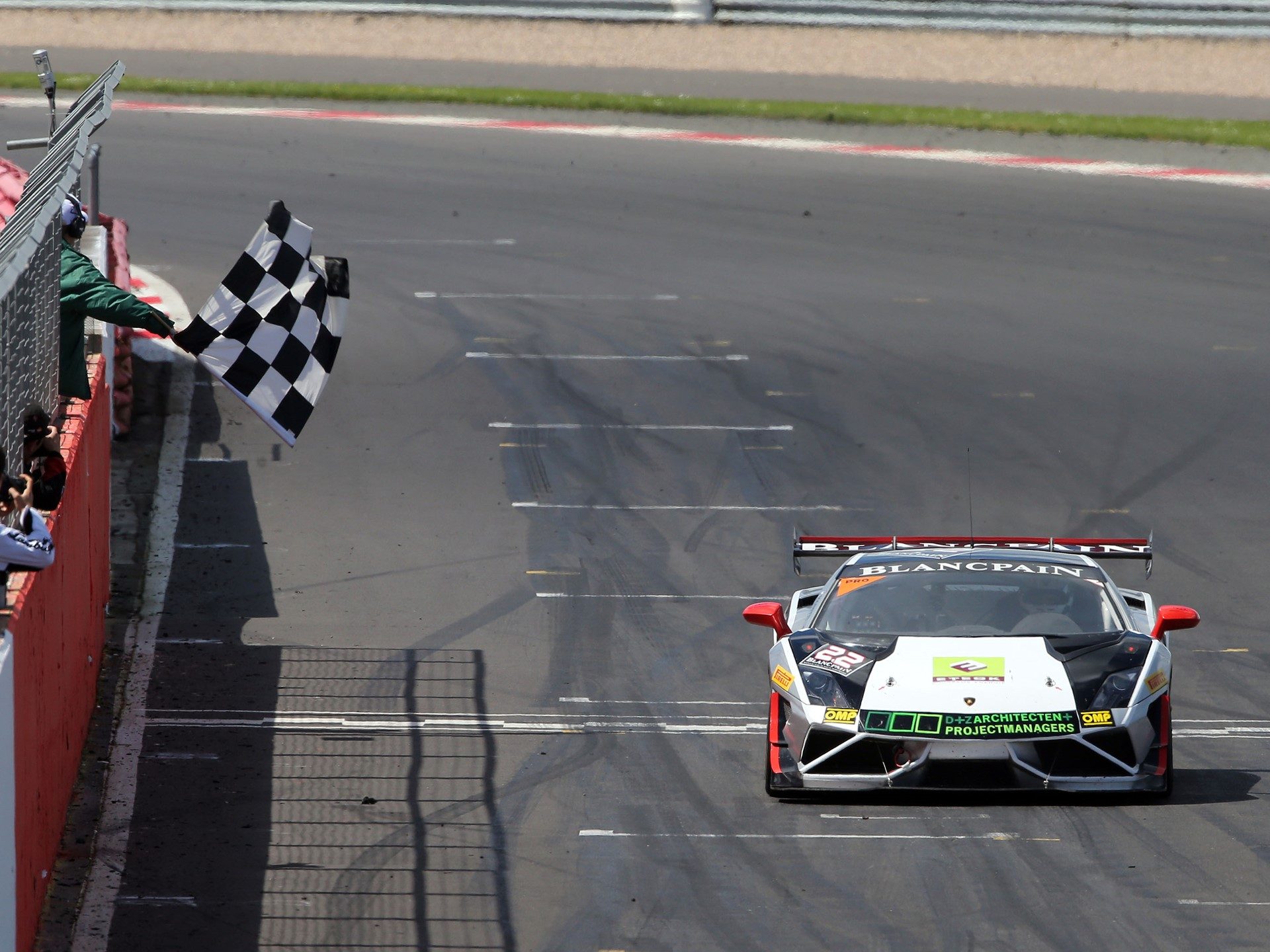 A Lamborghini Gallardo Super Trofeo race car crosses the finish line under a checkered flag at Silverstone