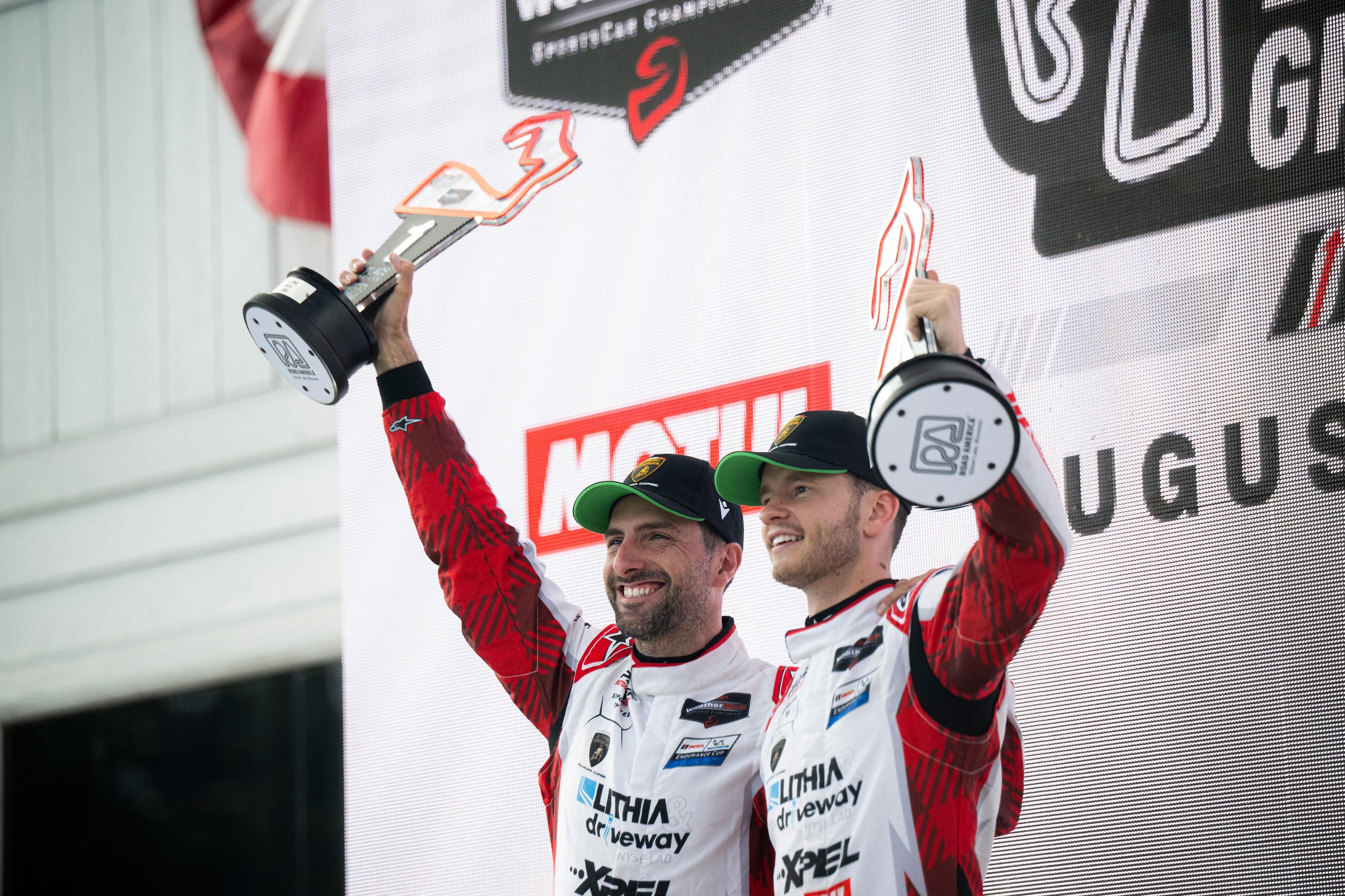 Temerario Gt3 Wins First Imsa Endurance Race At Road America, Five Months After Its Debut 7 Andrea caldarelli and sandy mitchell in red and white racing suits holding trophies on the road america podium