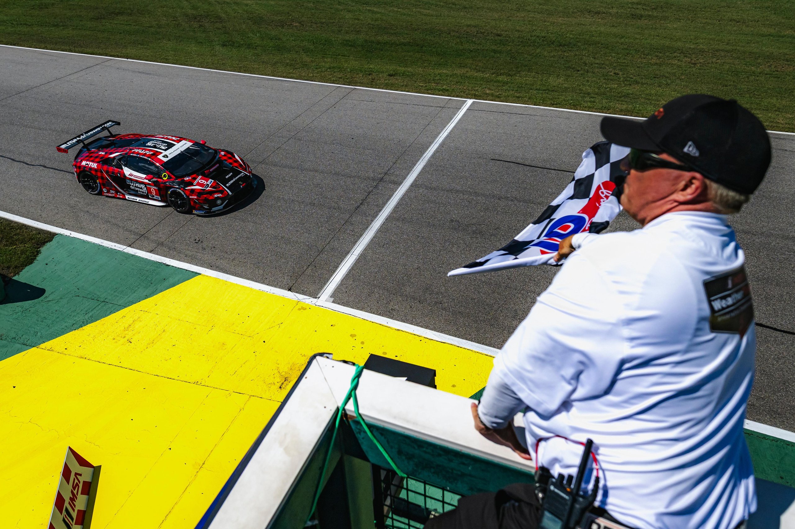 A red and black Lamborghini Huracán GT3 EVO2 race car with sponsor decals speeds past a race official waving a checkered flag from a high vantage point at a racetrack.