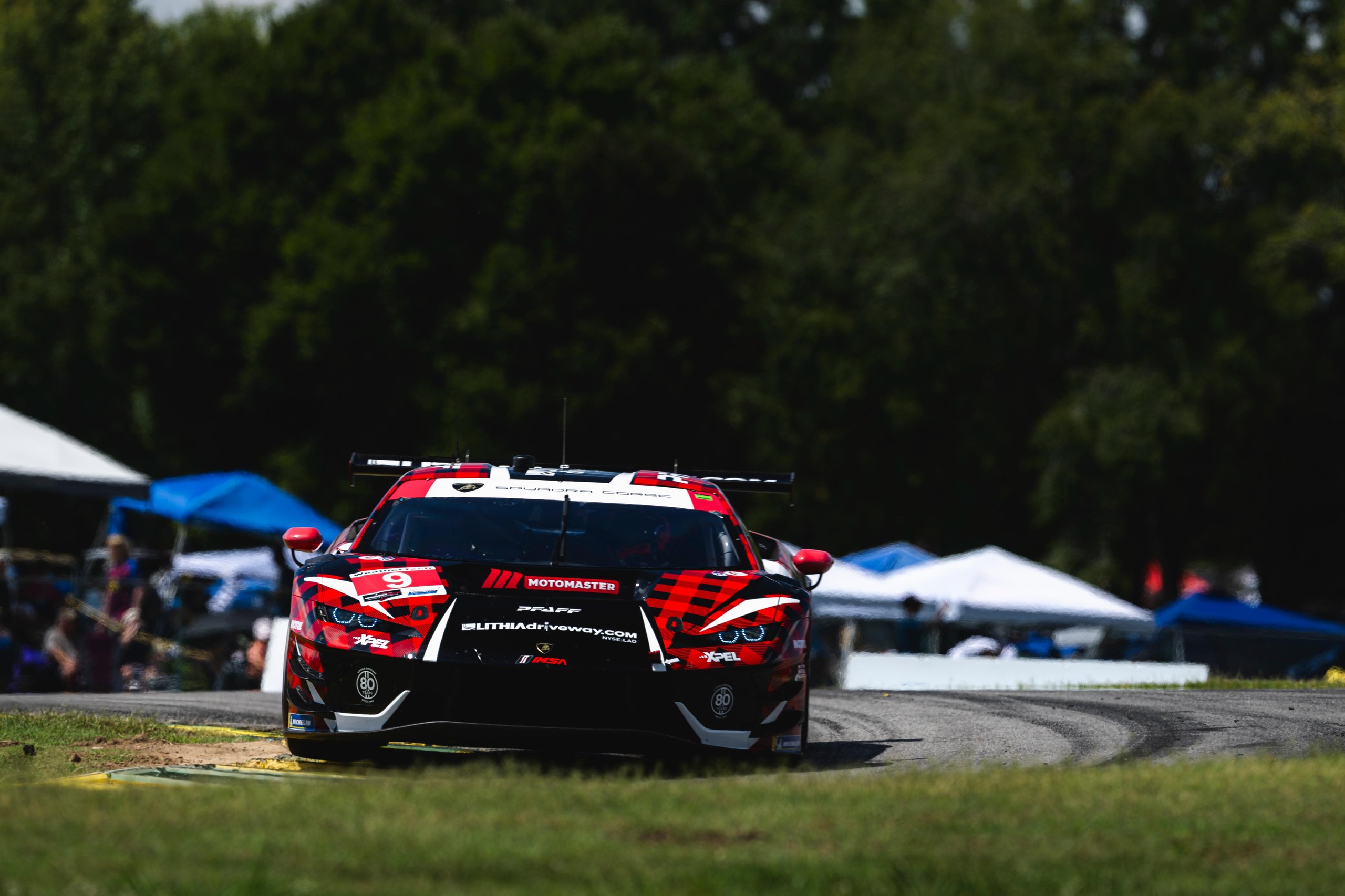 A red and black Lamborghini Huracán GT3 EVO2 race car, with a plaid-like livery and sponsor decals, approaches on a racetrack with blurred green trees in the background.