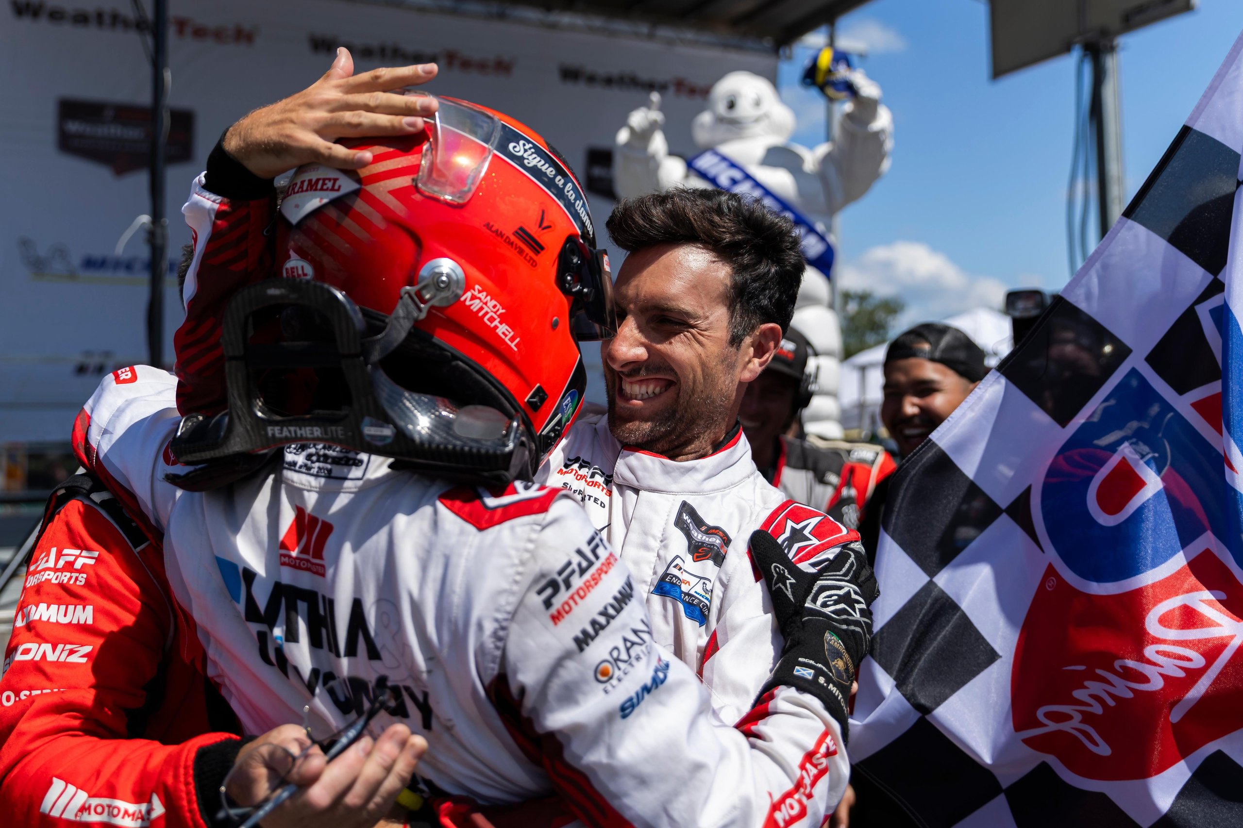 Two male race car drivers, one in a helmet and one without, embrace in celebration on a sunny day, with a checkered flag and a Michelin Man mascot in the background.