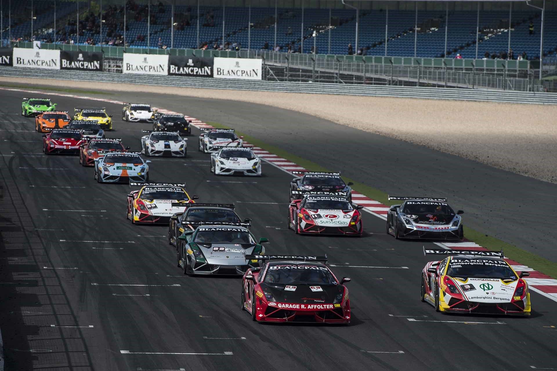 Pack of Lamborghini Gallardo Super Trofeo race cars battling for position on track with grandstands in the background