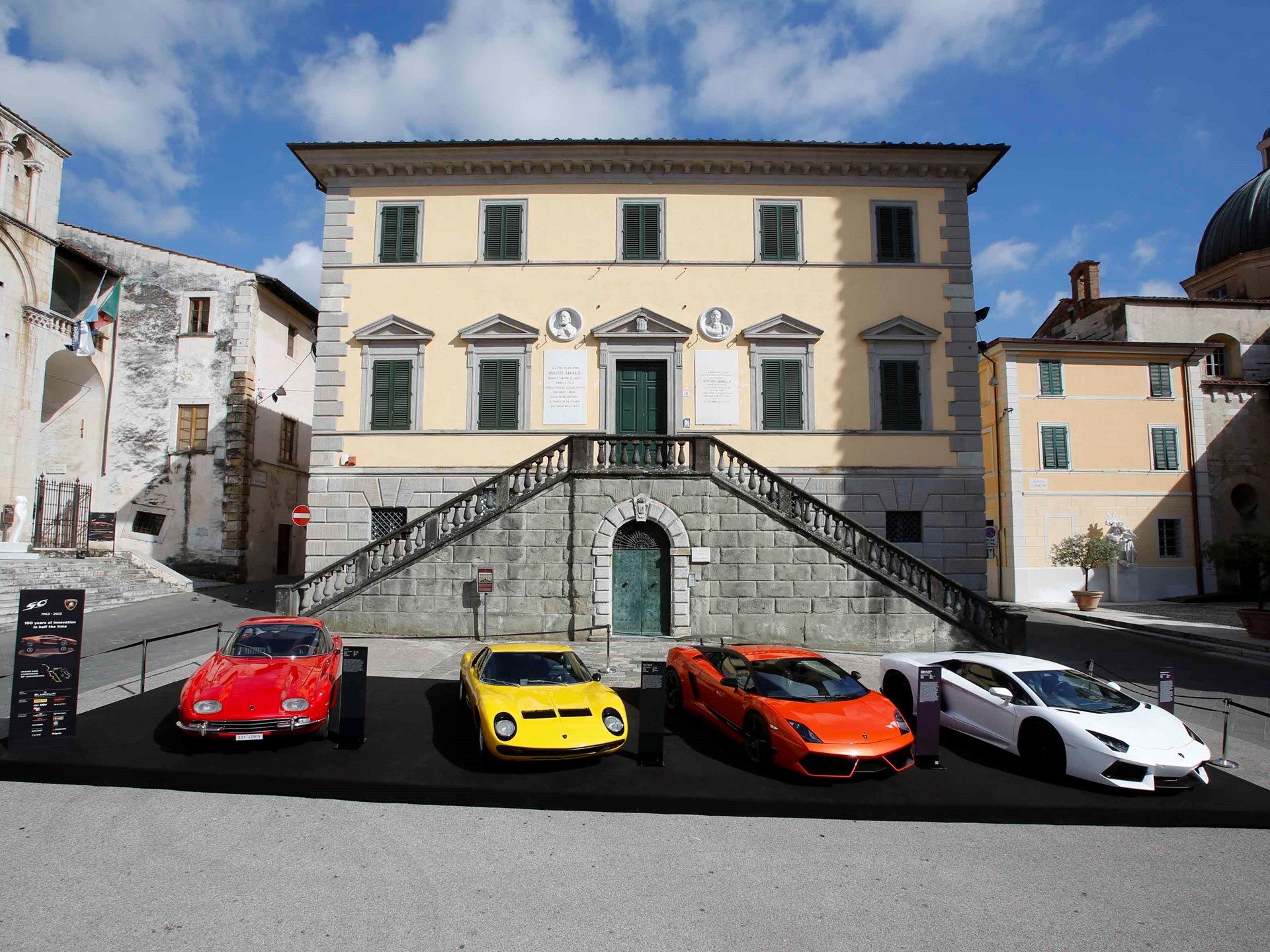 Four Lamborghini models spanning decades of design evolution, from the red 350 GT and yellow Miura to an orange Gallardo and white Aventador, displayed in front of a historic Tuscan building