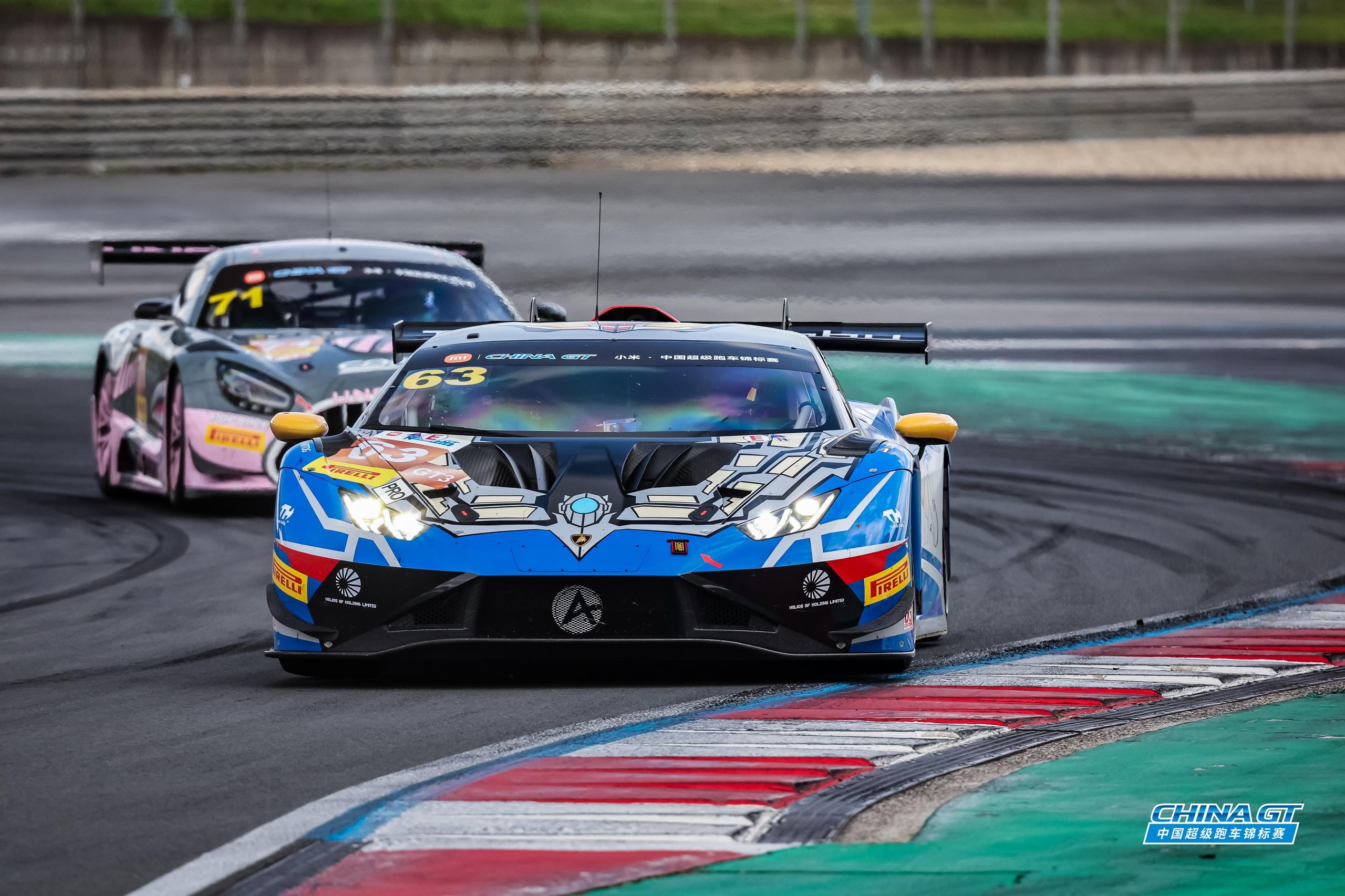 A blue and white Lamborghini Huracán GT3 EVO2 race car with number 63 leads another car on a racetrack, highlighting competitive racing.