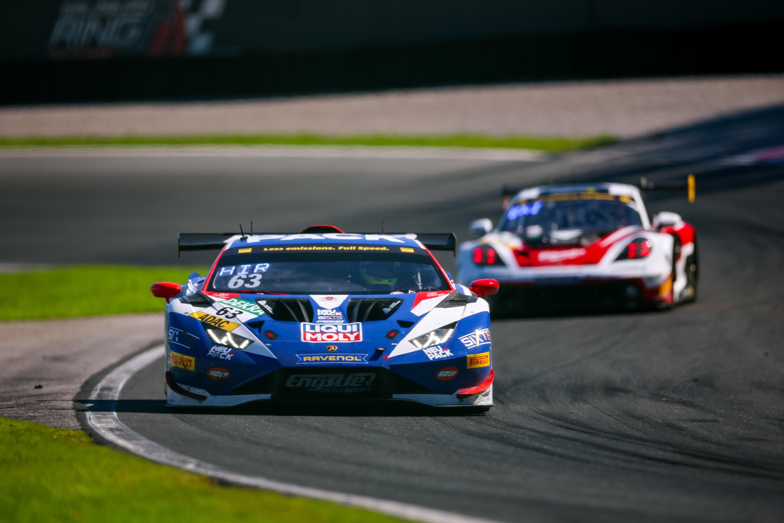 A blue, white, and red Lamborghini Huracán GT3 EVO2 race car with number 63 leads another car on a racetrack, capturing intense racing.