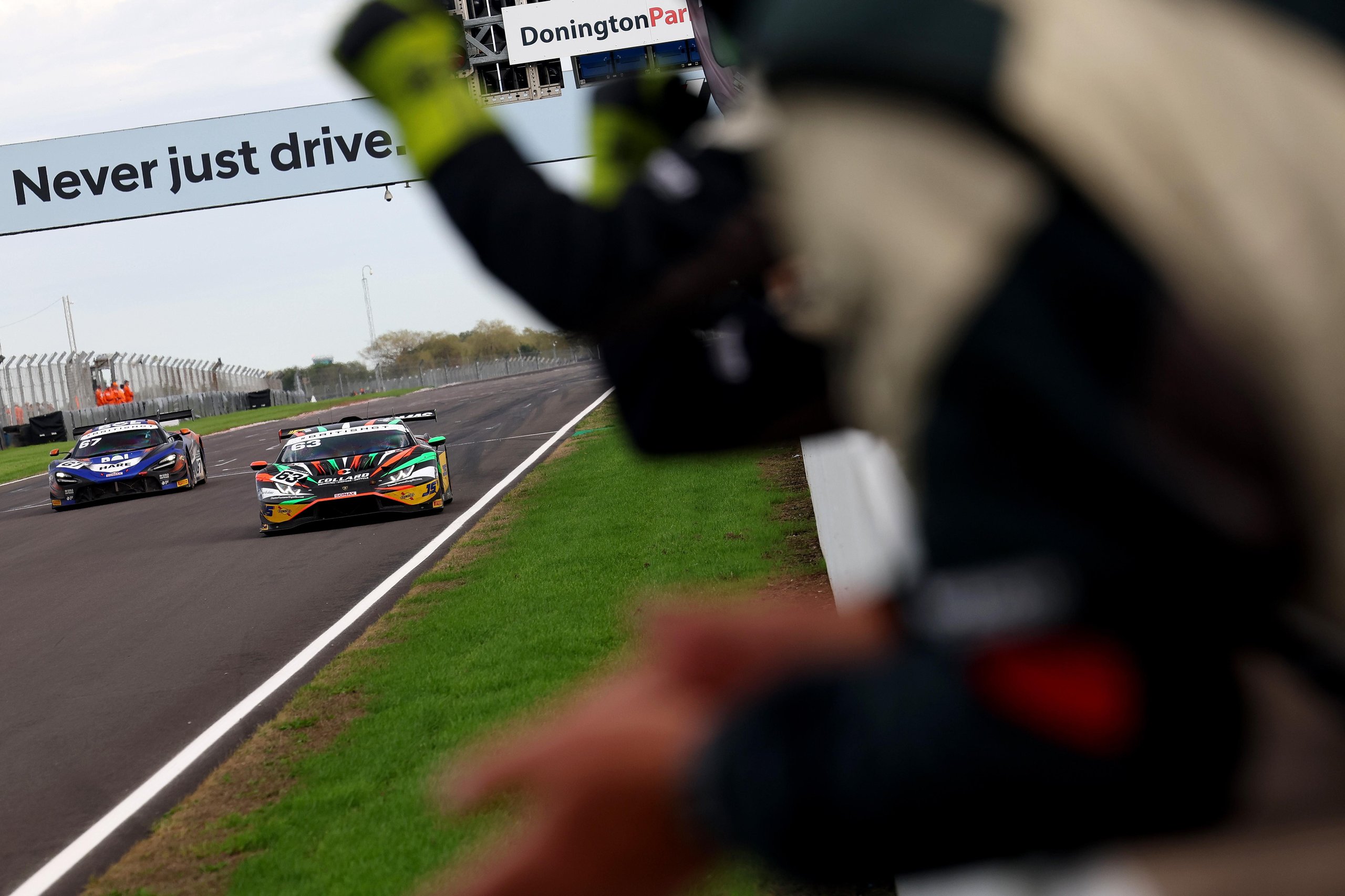 Two race cars, including a multi-colored Lamborghini Huracán GT3 EVO2, speed down a racetrack, viewed from behind a spectator.
