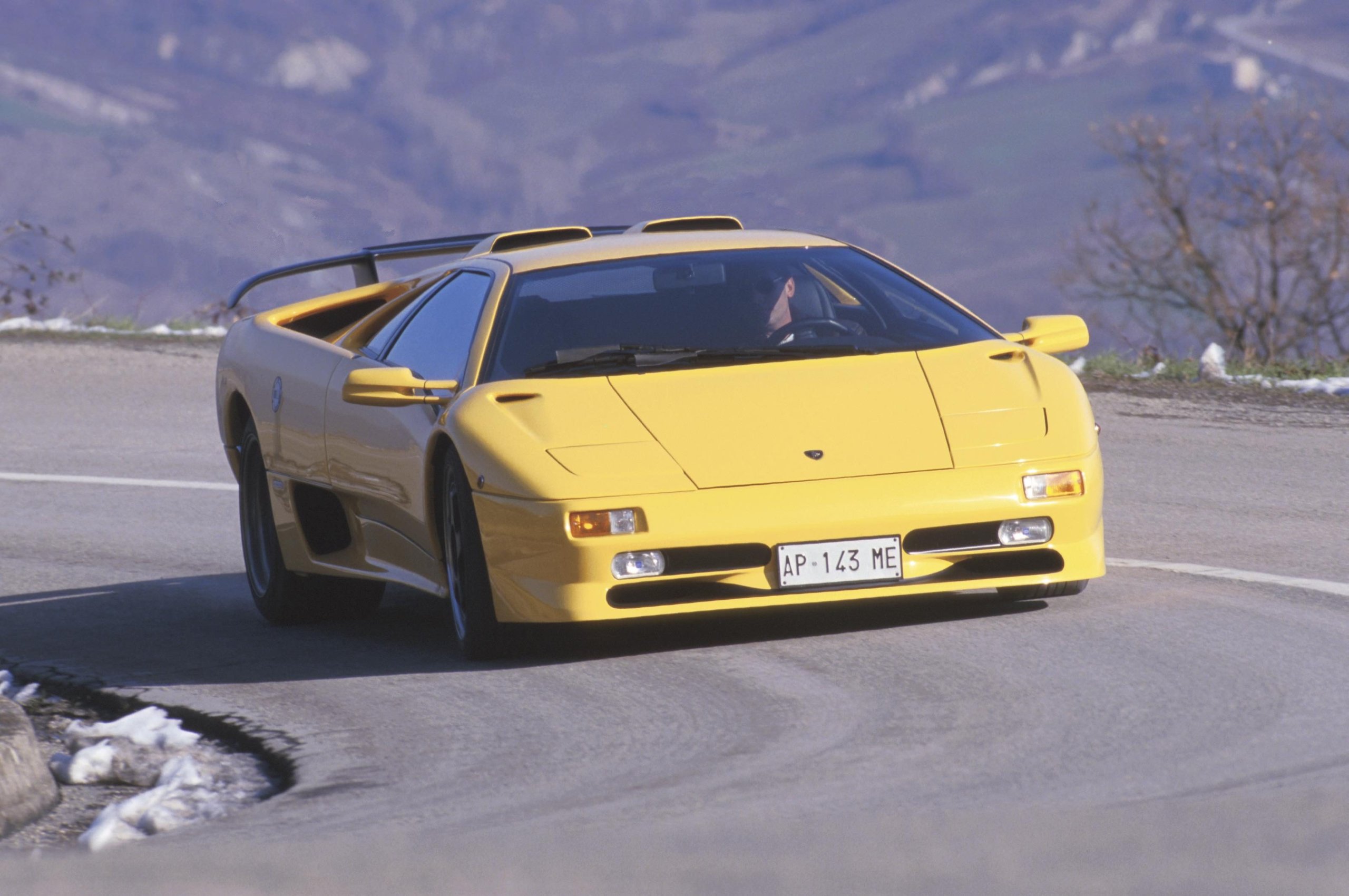 Yellow Lamborghini Diablo SV driving on a winding mountain road, showcasing its aggressive rear wing and classic supercar profile