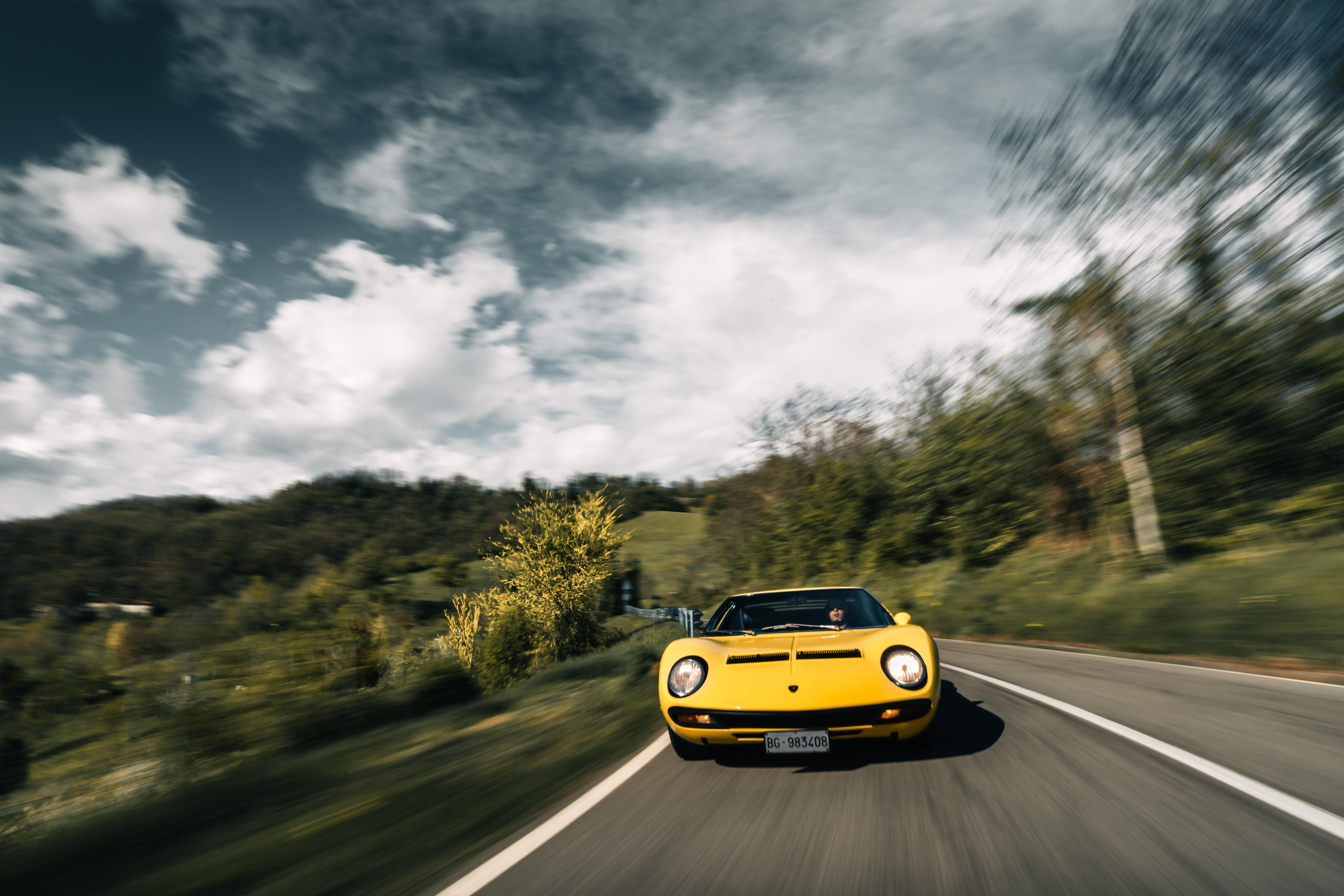 A bright yellow Lamborghini Miura SV is driving on a winding asphalt road through a lush green, hilly landscape. The car is angled slightly towards the viewer, with motion blur indicating speed. A dramatic sky with white and grey clouds is above. The license plate 'BG - 983408' is visible on the fro