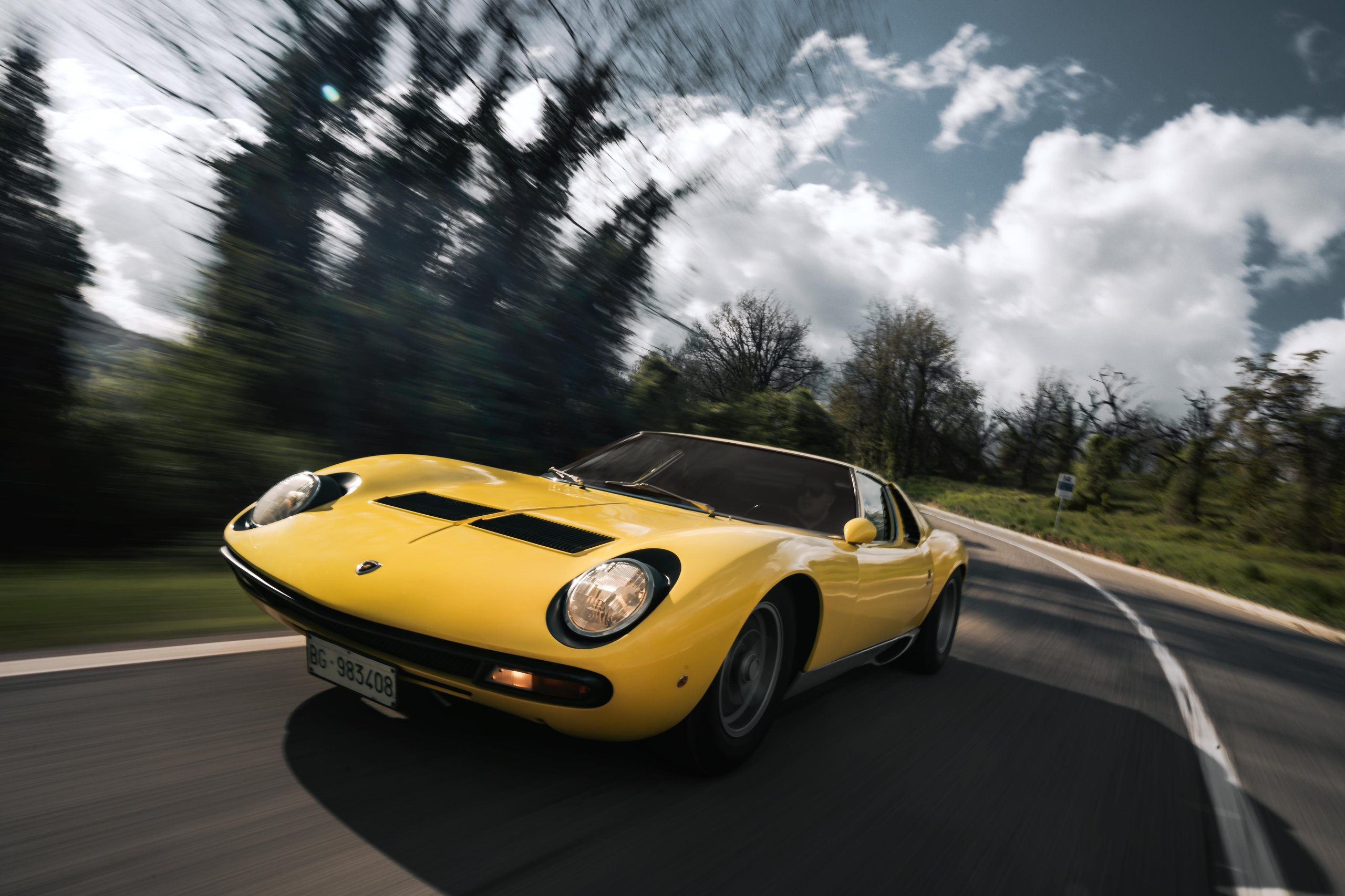 A yellow Lamborghini Miura SV is captured in a dynamic action shot, speeding along a winding road with a motion-blurred background of trees and sky.