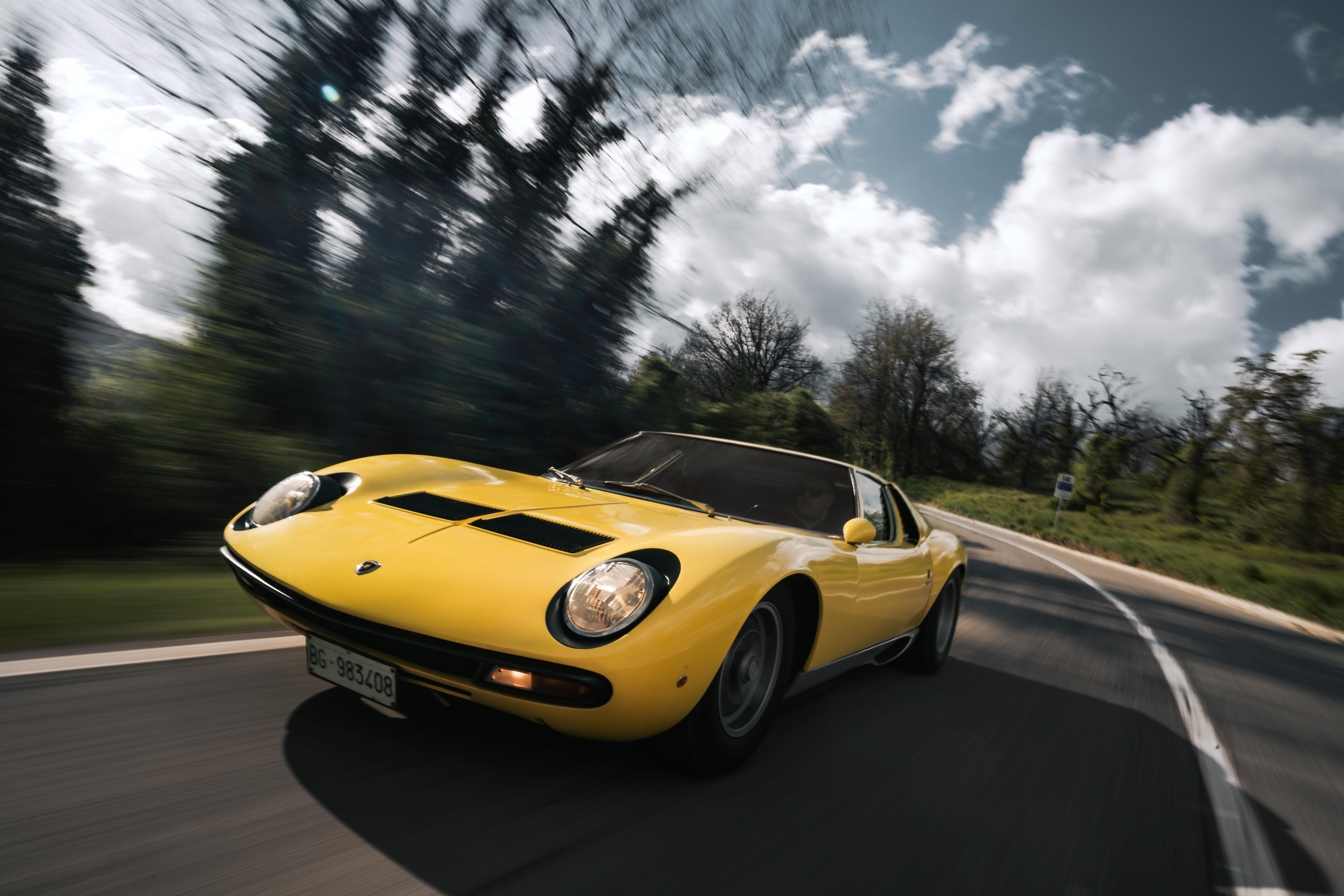 A yellow Lamborghini Miura SV is captured in a dynamic action shot, speeding along a winding road with a motion-blurred background of trees and sky.