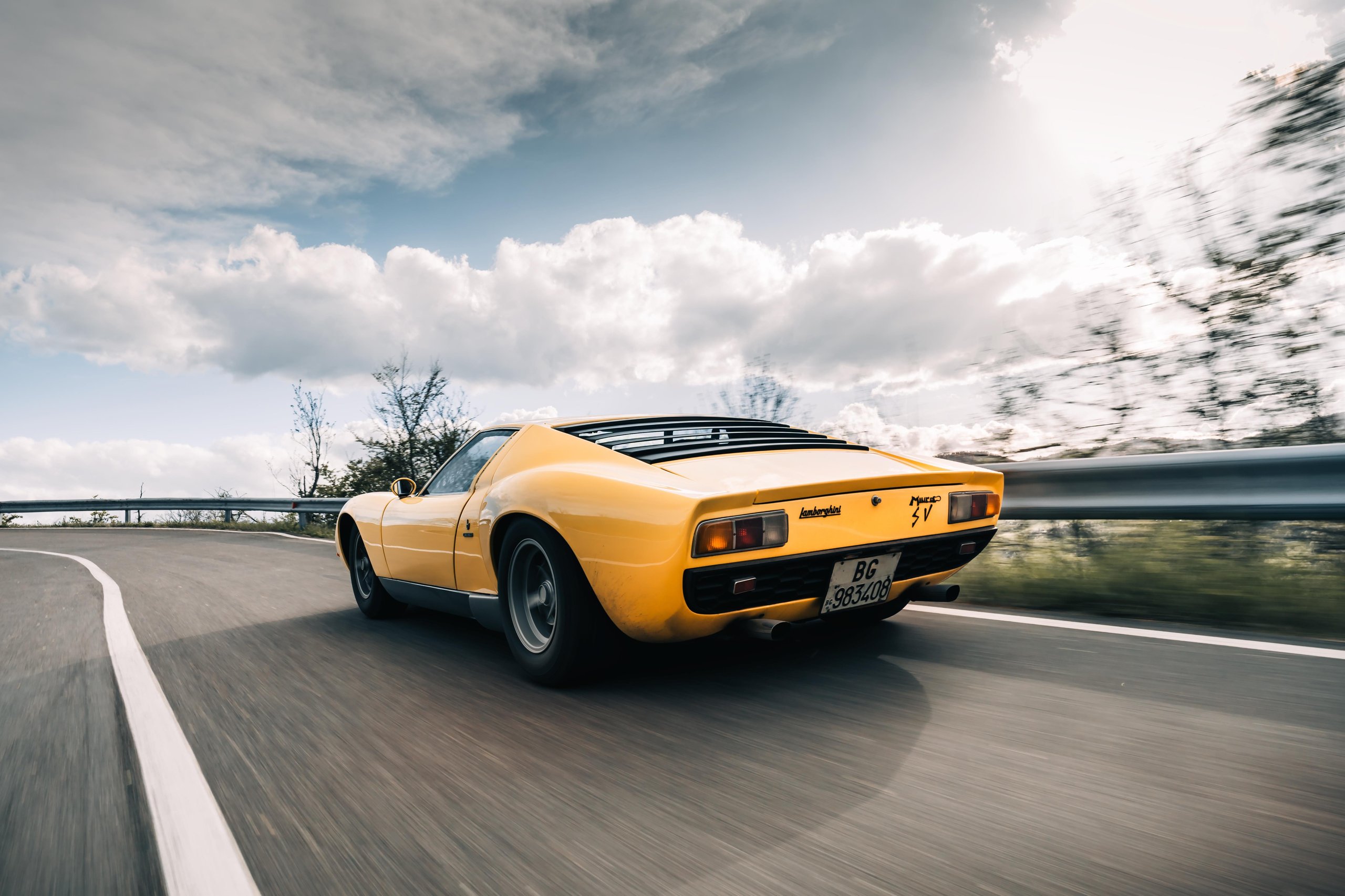 A yellow Lamborghini Miura SV is seen from the rear-three-quarter view, driving on a winding road with a motion-blurred background of trees and a cloudy sky.