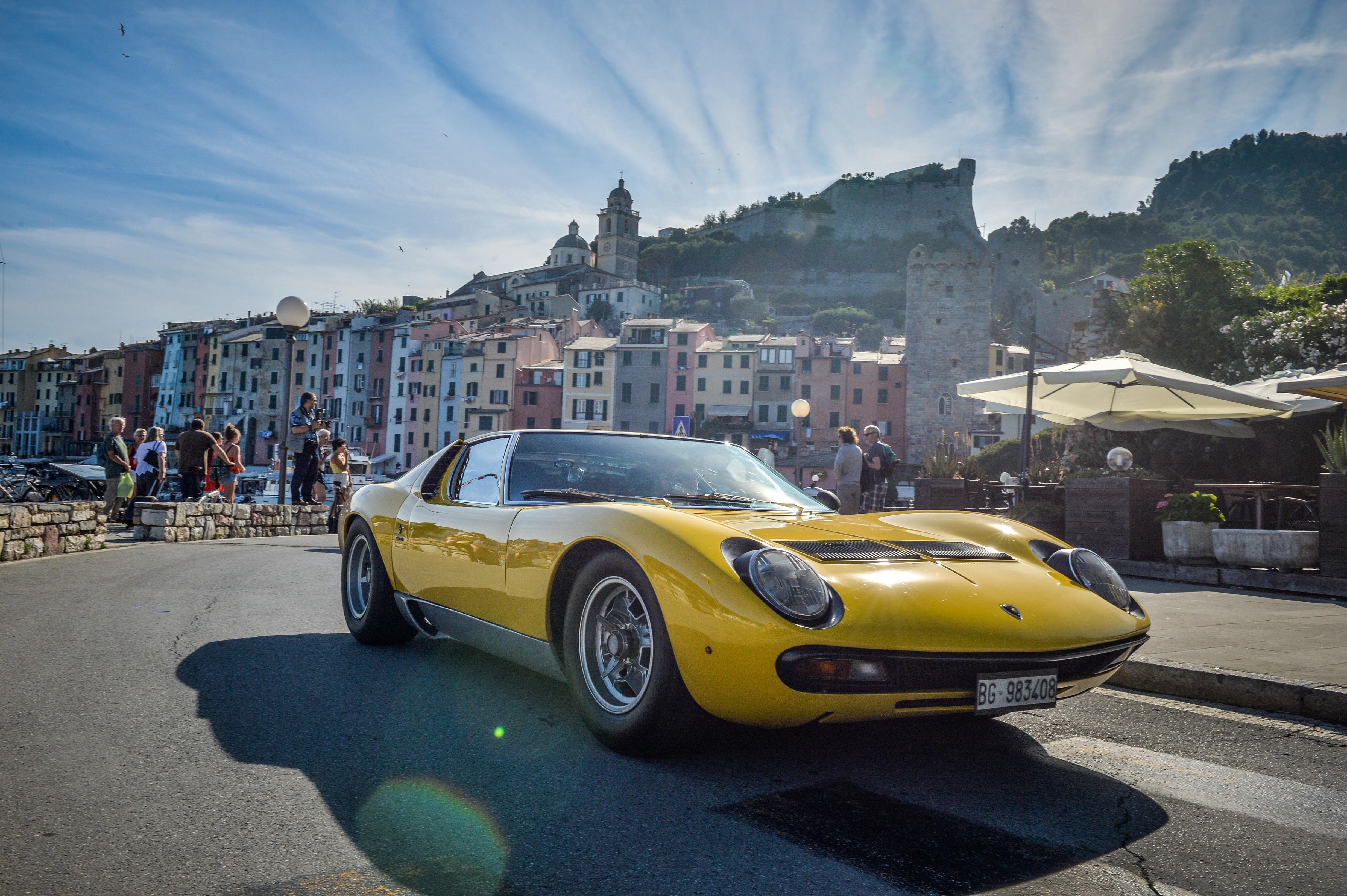 A bright yellow Lamborghini Miura SV is parked on the side of a street in a picturesque coastal town. Colorful buildings line the street, and a historic castle or fortress is visible on a hill in the background under a blue sky with white clouds. Several people are walking in the background. The car