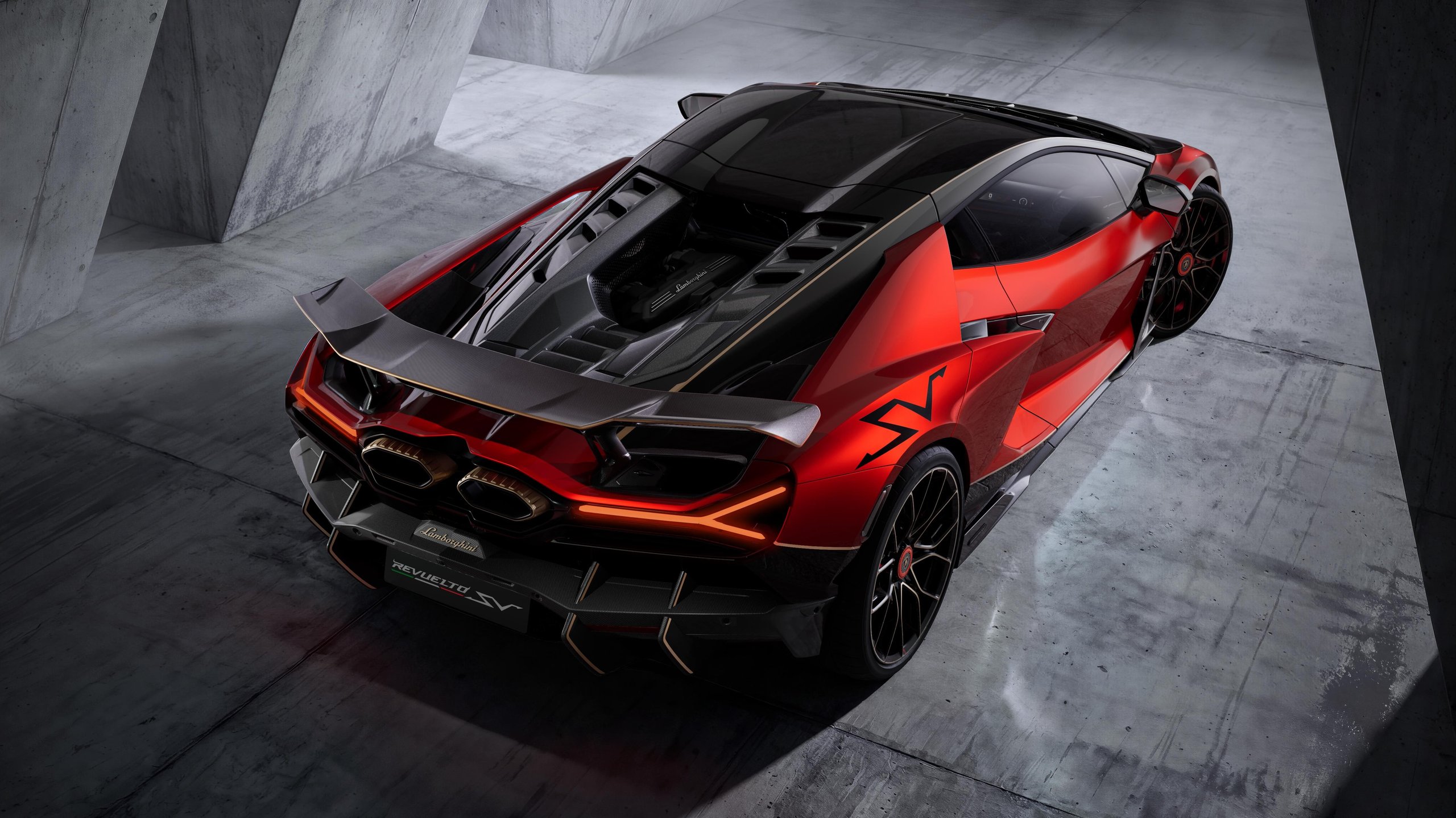 A red and black Lamborghini Revuelto SV is shown from a high rear-side angle in a studio with concrete walls. The exposed engine, large rear wing, and distinctive exhaust outlets are prominent.