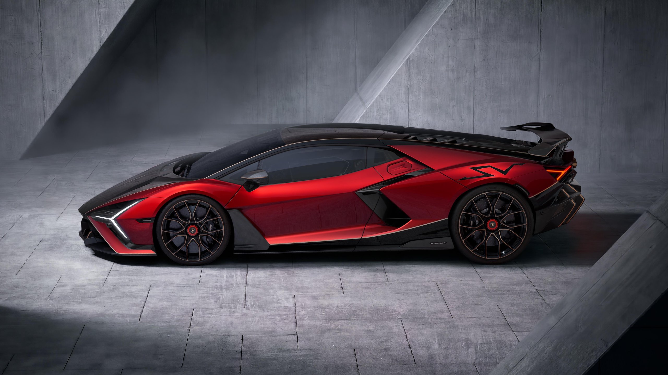 A red and black Lamborghini Revuelto SV is shown from a side profile in a studio with concrete walls. The car's aerodynamic silhouette, large black wheels, and distinctive side vents are clearly visible.